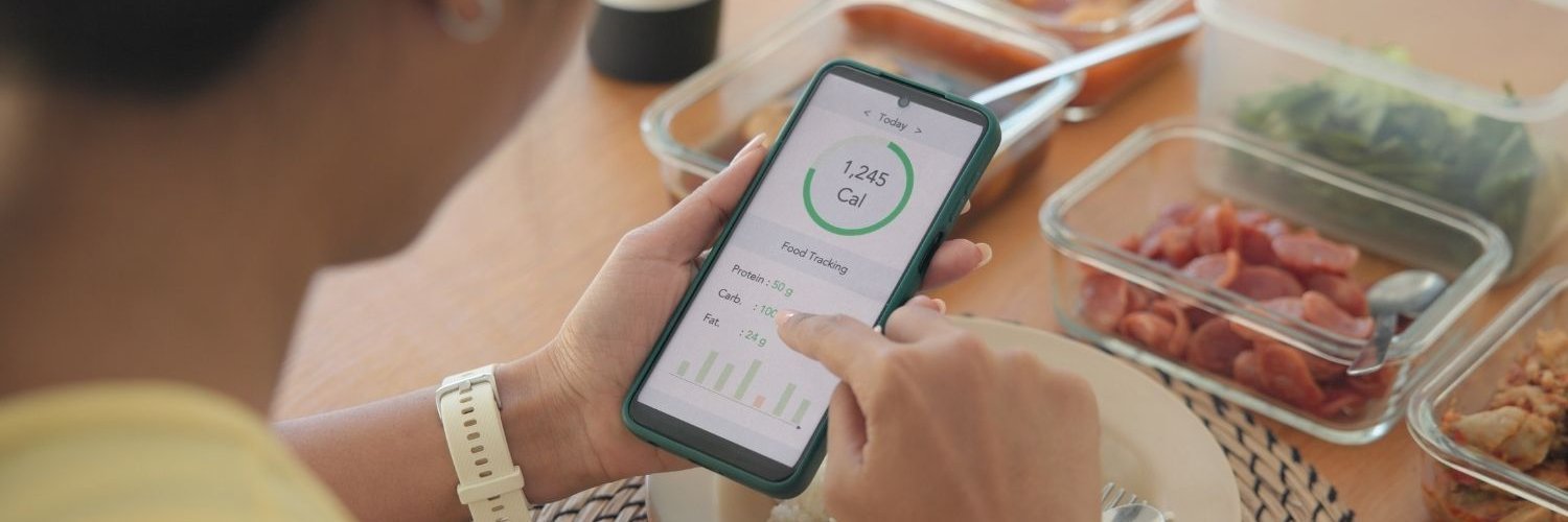 Eine junge Frau sitzt am Tisch und schaut auf ihr Smartphone, wo Kalorien und Nährwerte angezeigt werden. Vor ihr stehen Behälter mit verschiedenen Gemüsesorten.