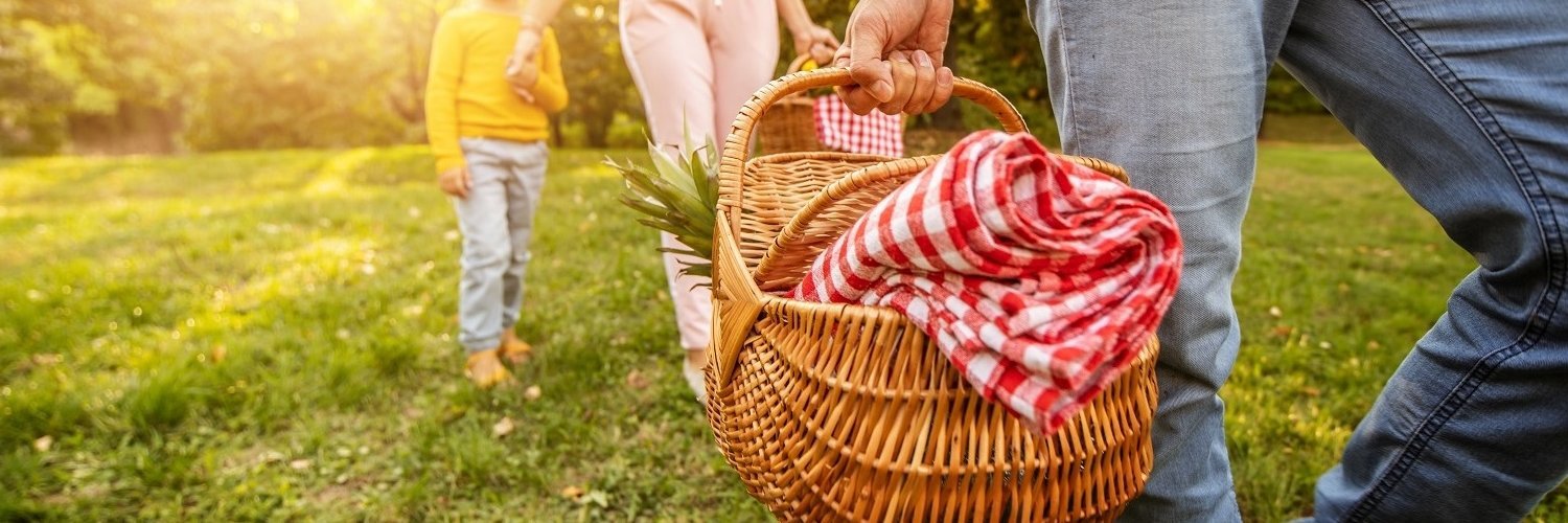 Ein Mann geht über eine Wiese und trägt einen Picknickkorb. Hinter ihm her laufen eine Frau und ein Kind. 