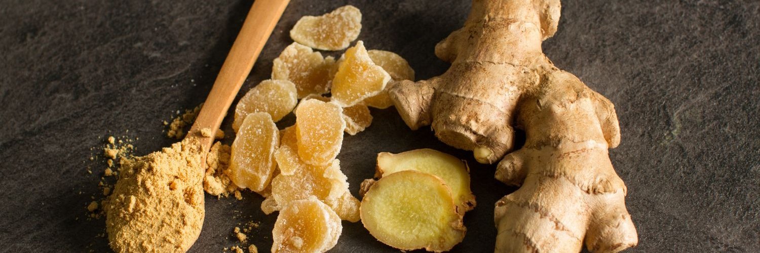 Eine Ingwerwurzel, kandierter Ingwer und Ingwerpulver in einem Holzlöffel liegen auf grauem Betonhintergrund.