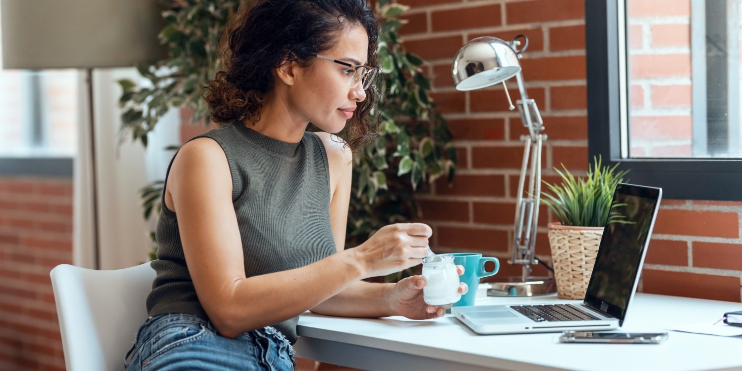 Eine Frau sitzt im Homeoffice vor ihrem Laptop und isst einen Joghurt. . Artikel "Fit und fokussiert im Homeoffice" öffnen.
