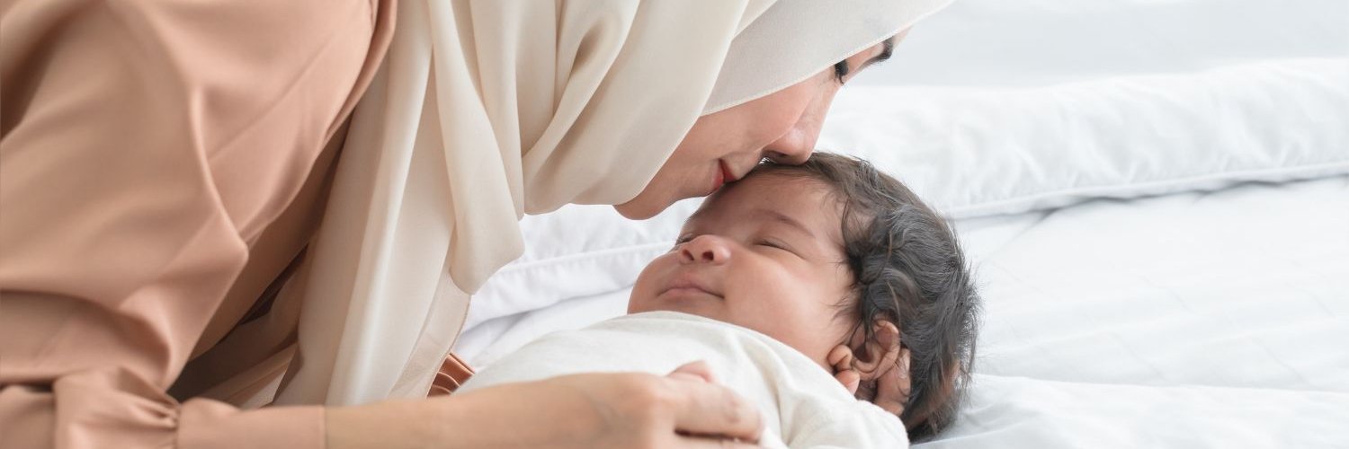 Eine junge Muslimin mit Hijab küsst ihr lächelndes, einmonatiges Neugeborenes auf die Stirn, während das Baby im Bett schläft.