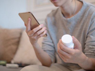 Eine junge Frau hält ein Smartphone in der einen Hand und liest. In der anderen Hand hält sie eine Nahrungsergänzungsmittel-Packung.