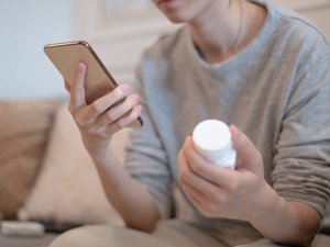Eine junge Frau hält ein Smartphone in der einen Hand und liest. In der anderen Hand hält sie eine Nahrungsergänzungsmittel-Packung.