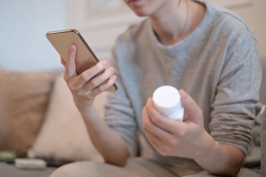 Eine junge Frau hält ein Smartphone in der einen Hand und liest. In der anderen Hand hält sie eine Nahrungsergänzungsmittel-Packung.