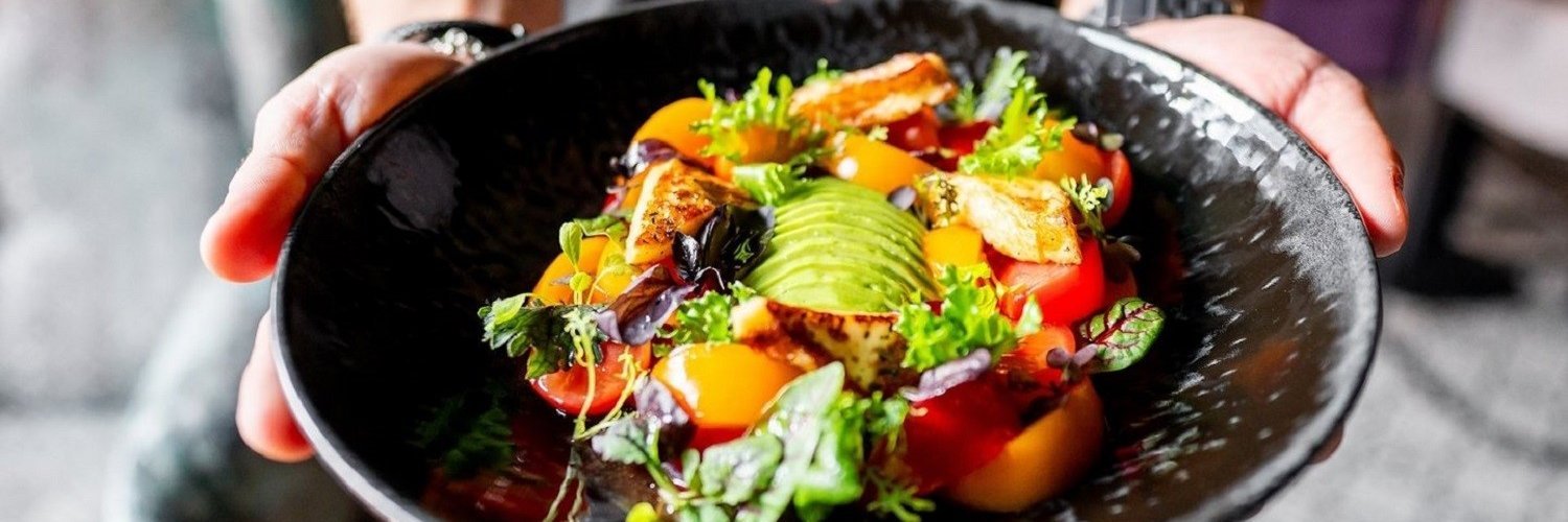 Eine Person hält einen schwarzen Teller mit einem bunten Salat aus Gemüse, Tomaten, Avocado und Tofu in der Hand. 