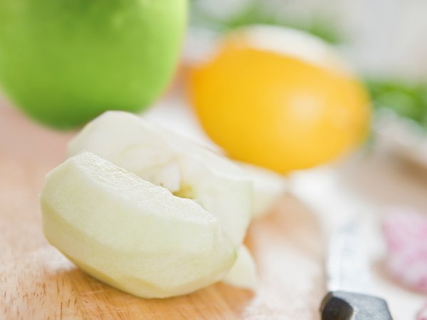 Auf einem Brett liegen ein halber geschälter Apfel, ein grüner Apfel, eine Zitrone und ein Küchenmesser.