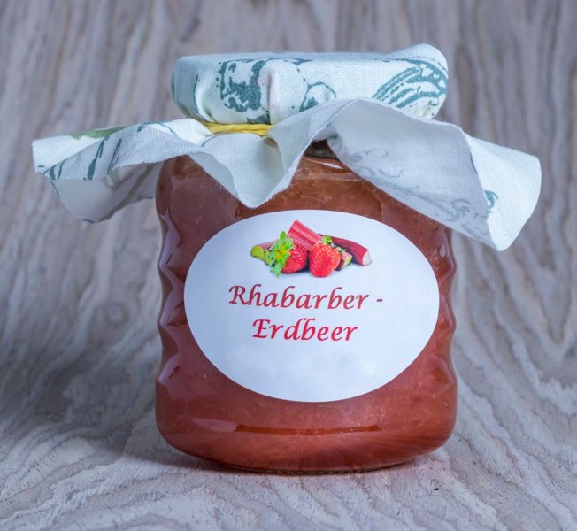 Ein Glas selbstgemachte Erdbeer-Rhabarber-Marmelade steht auf hellgrauem Hintergrund.