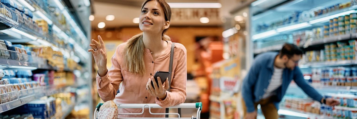 Eine Frau beim Einkaufen im Supermarkt, sie schiebt einen Einkaufswagen und hält ihr Handy in der Hand.