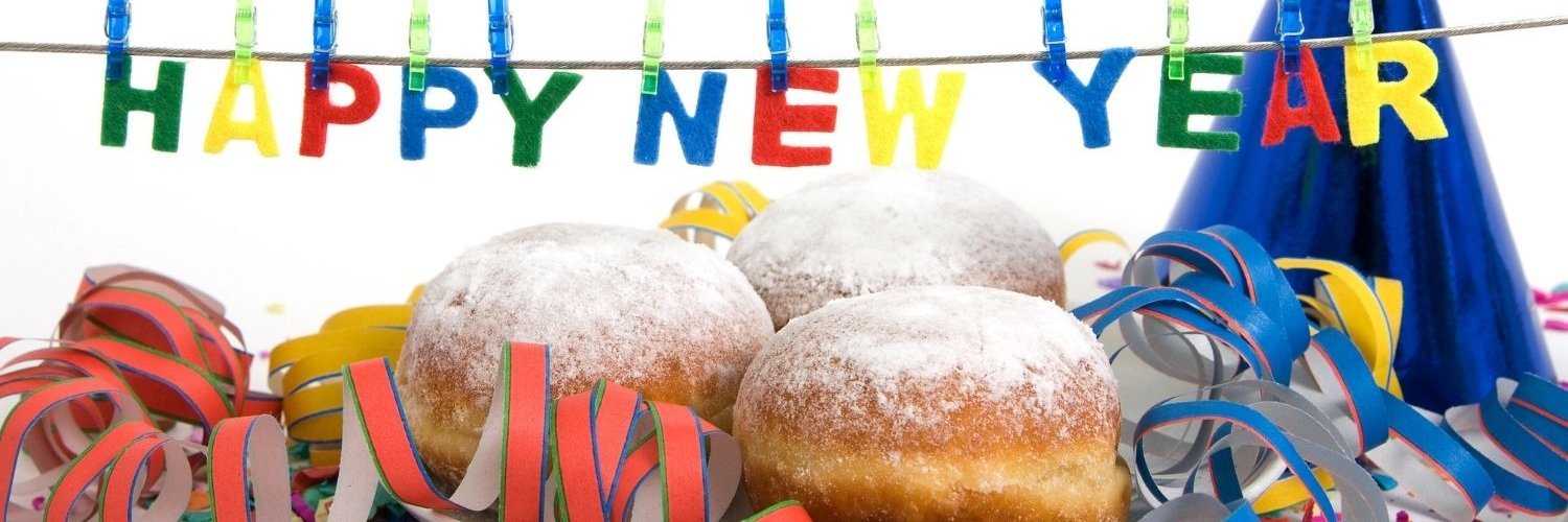 Auf einem Tisch steht ein Teller mit Berlinern, dekoriert mit Luftschlangen und darüber hängt eine Schnur mit Buchstaben an Wäscheklammern, die „Happy New Year“ formen.