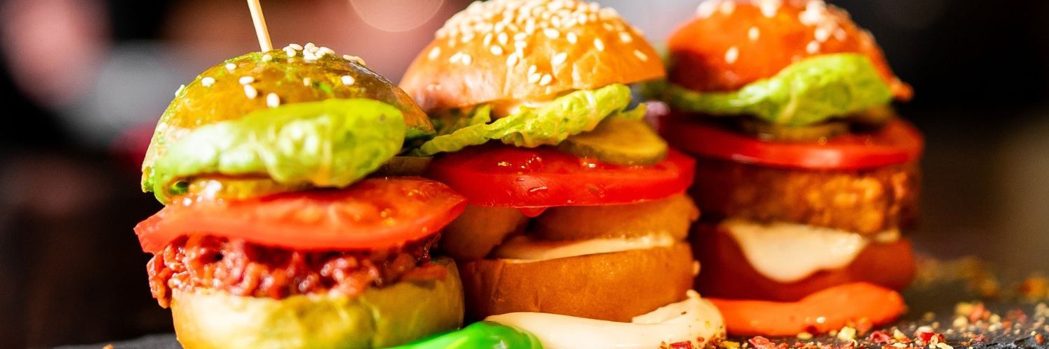 Auf einer dunklen Fläche stehen drei Miniburger mit verschiedenen Zutaten gefüllt und jeweils einem Holzspieß fixiert.