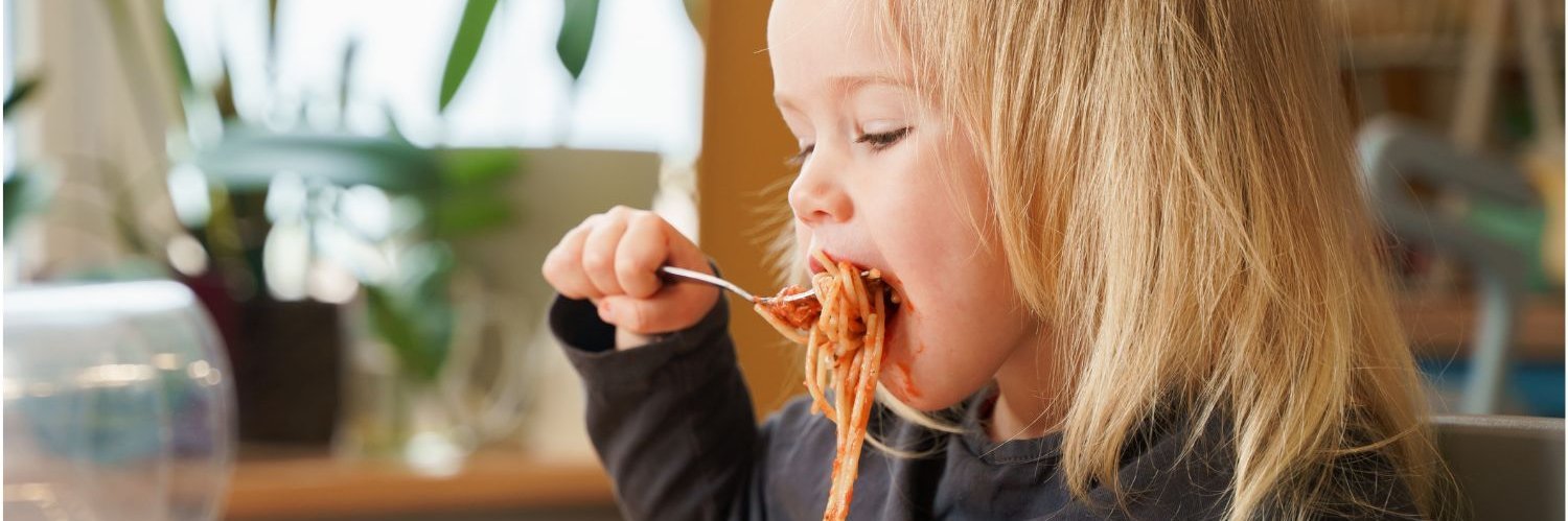 Ein blondes Mädchen im Kleinkindalter sitzt an einem Esstisch und schiebt sich eine volle Gabel mit Spaghetti in den Mund.