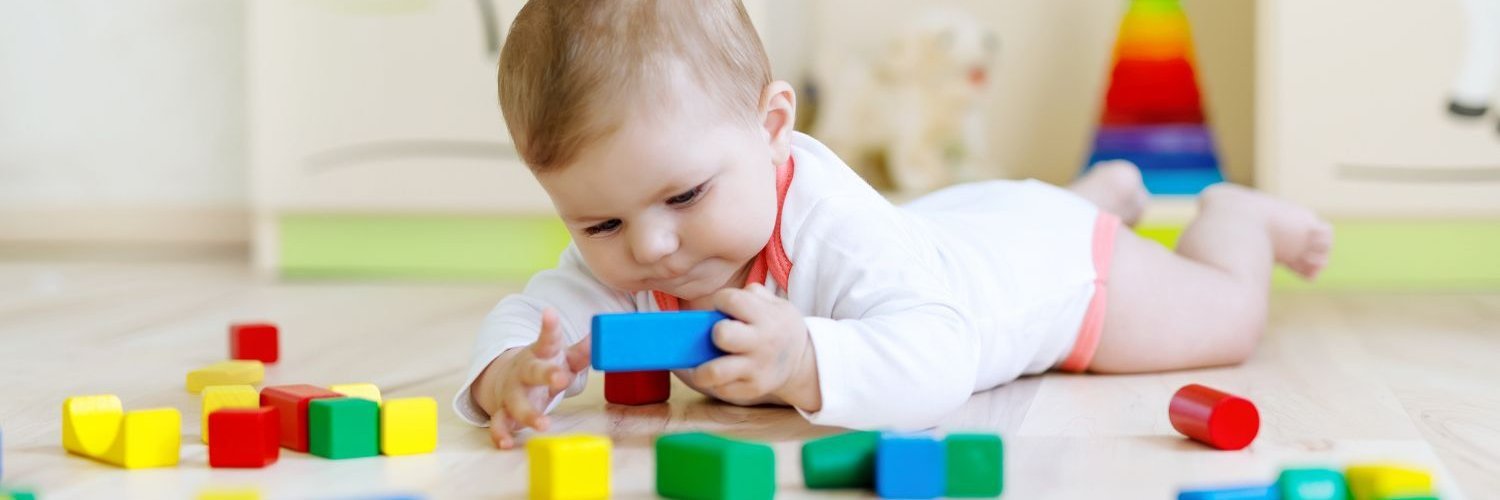Ein Baby liegt auf dem Bauch auf dem Fußboden und spielt mit bunten Bauklötzen. 