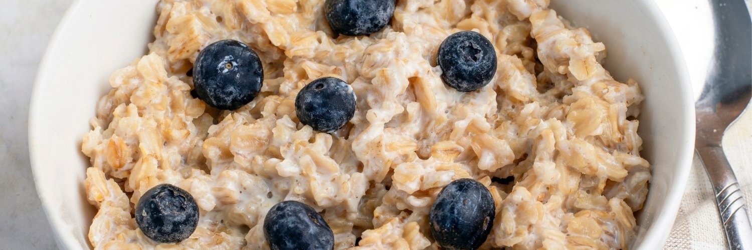 In einer weißen Schüssel sind Haferflocken als Porridge angerichtet und mit Blaubeeren garniert, daneben liegt ein Löffel.