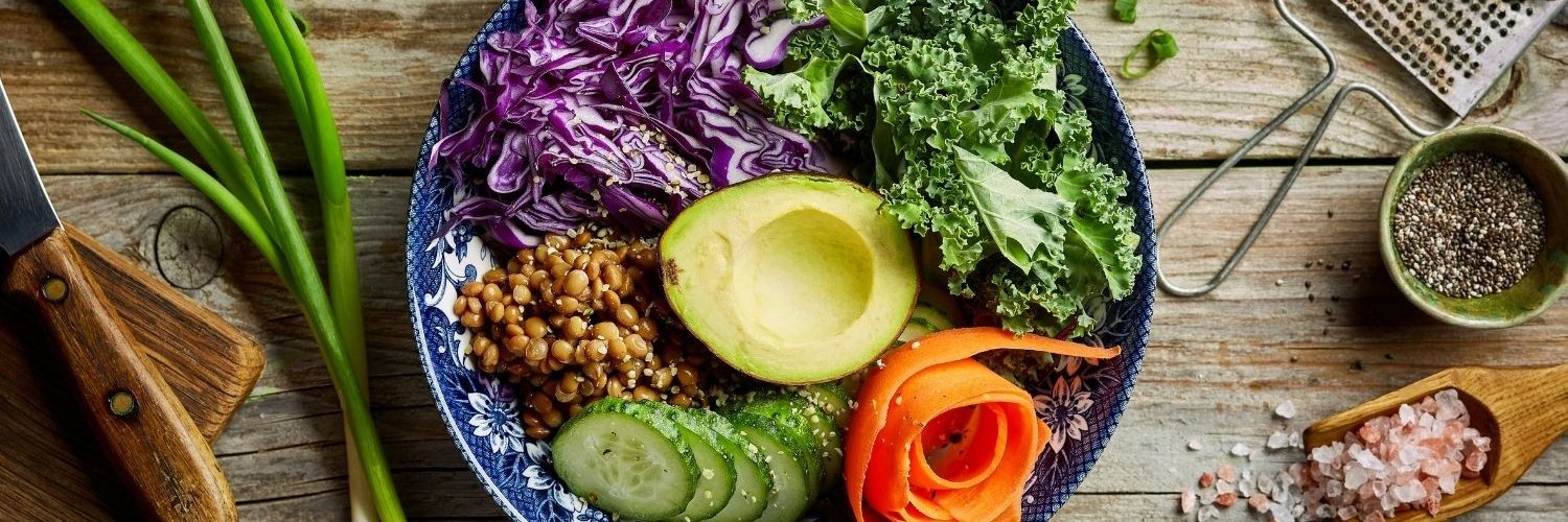 In einer blau-weiß gemusterten Schüssel sind Rotkohlstreifen, Blattsalat, Linsen, Salatgurke, Möhre und Avocado angerichtet. Auf dem Tisch daneben liegen Frühlingszwiebeln, Salz, Pfeffer und Muskatnuss.