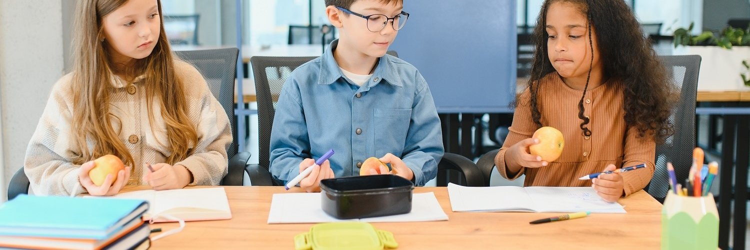 Zwei Mädchen und ein Junge sitzen in der Schule an einem Tisch im Klassenraum. Sie haben ihre Hefte geöffnet, jeweils einen Apfel in der Hand und schauen konzentriert. 