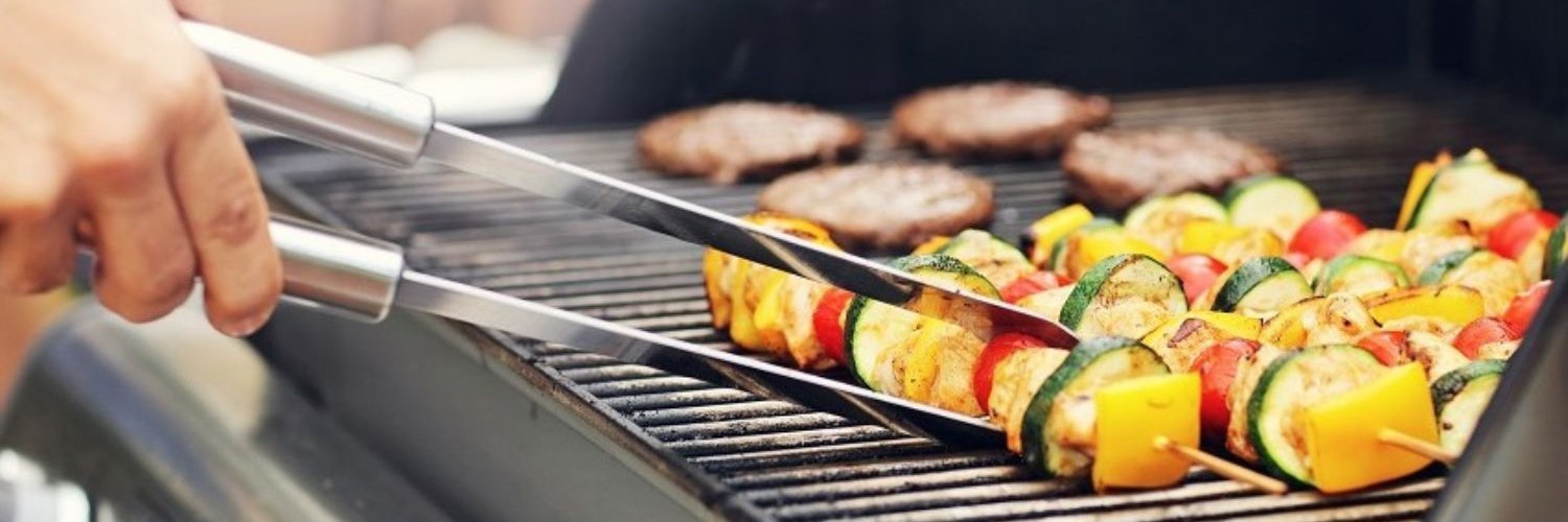 Auf einem Grillrost eines Gasgrills liegen Gemüsespieße und Fleisch-Patties. Ein Spieß wird mit einer Grillzange gewendet.
