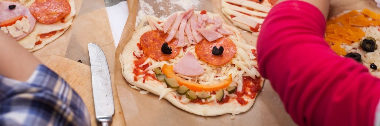Kinder belegen ausgerollte Teigfladen mit verschiedenen Pizzazutaten und bilden daraus lustige Gesichter. 