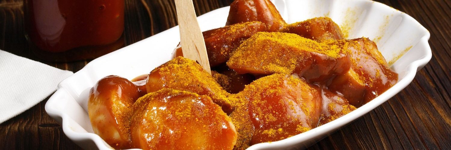 Auf einem dunklen Holztisch steht eine weiße Porzellanschale mit in Stücke geschnittener Currywurst mit Currypulver bestäubt. Daneben steht Curry-Ketchup.