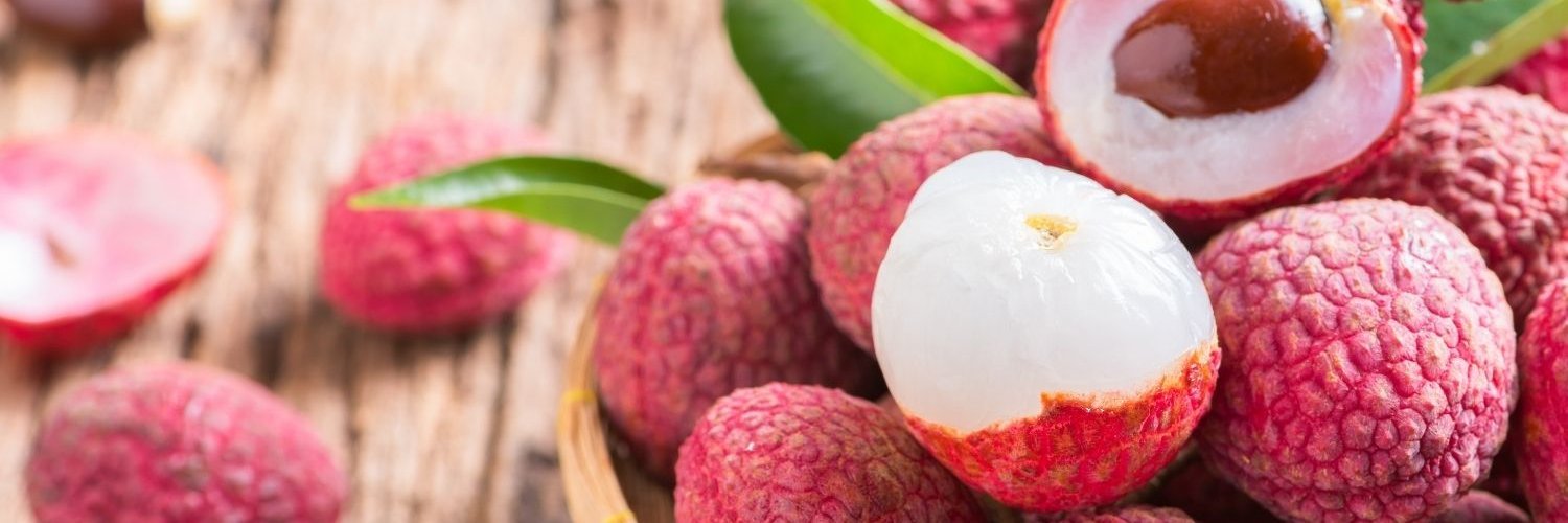 In einer Holzschale liegen Litschi teils mit rosafarbener Schale und teils geschält und halbiert, sowie einige schmale grüne Blätter. 