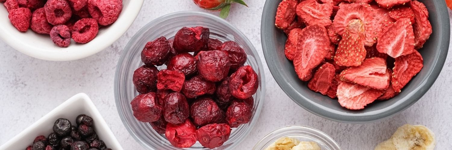Auf einem weißen Untergrund stehen mehrere Schüsseln mit gefriergetrockneten Himbeeren, Erdbeeren, Kirschen, Blaubeeren und Bananen.
