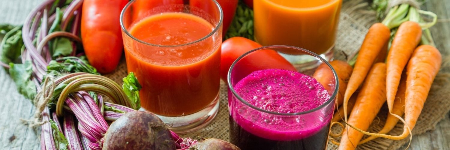 Auf einem Holztisch stehen drei Gläser mit Rote-Bete-, Möhren- und Tomatensaft. Daneben liegen Tomaten, Möhren und Rote-Bete-Knollen.