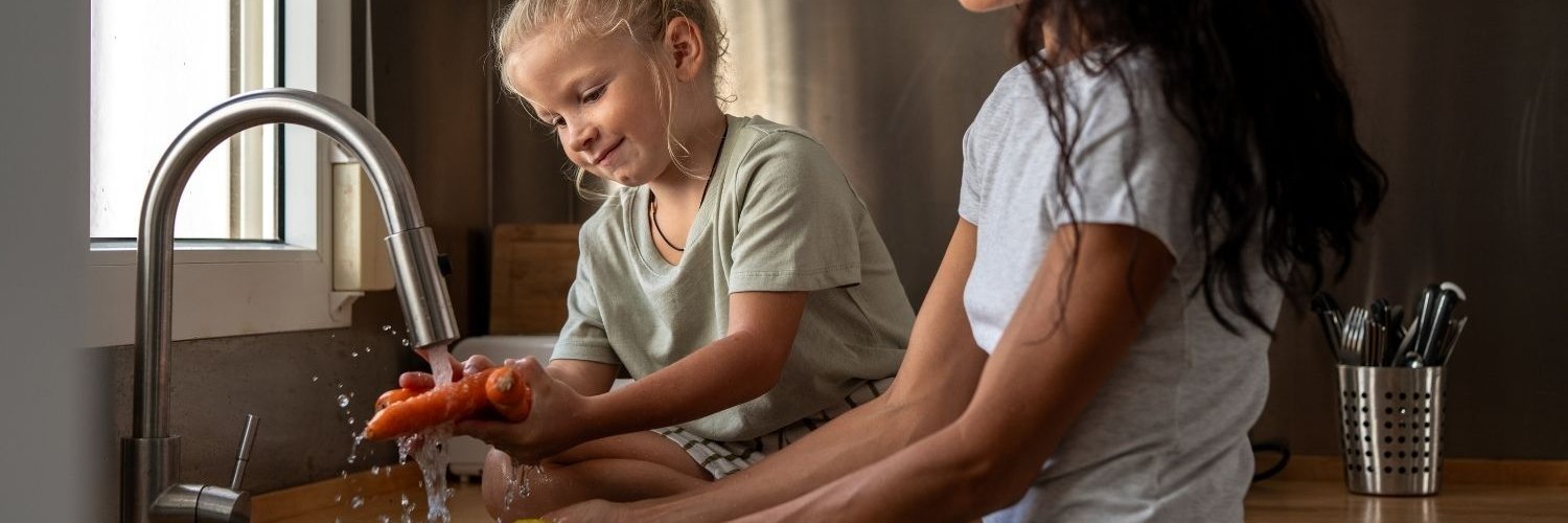 Eine dunkelhaarige Frau und ein Kind waschen in einer Küchenspüle Möhren und Äpfel unter fließendem Wasser.