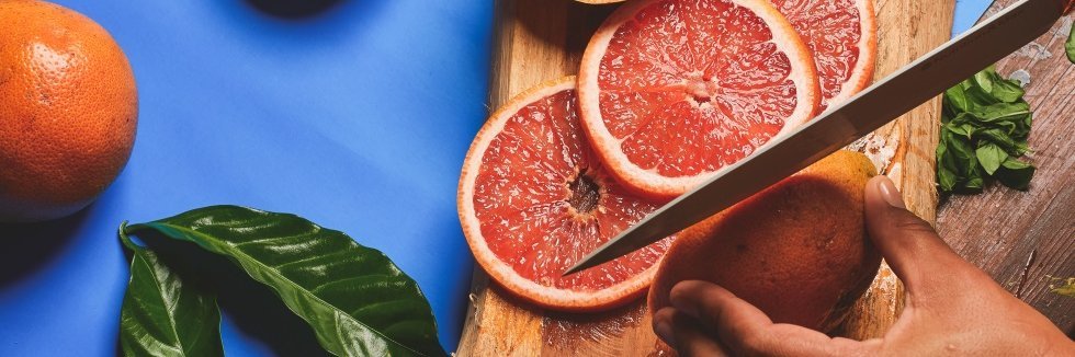 Zwei Hände halten eine Grapefruit auf einem Holzbrett fest und schneiden sie in dünne Scheiben.