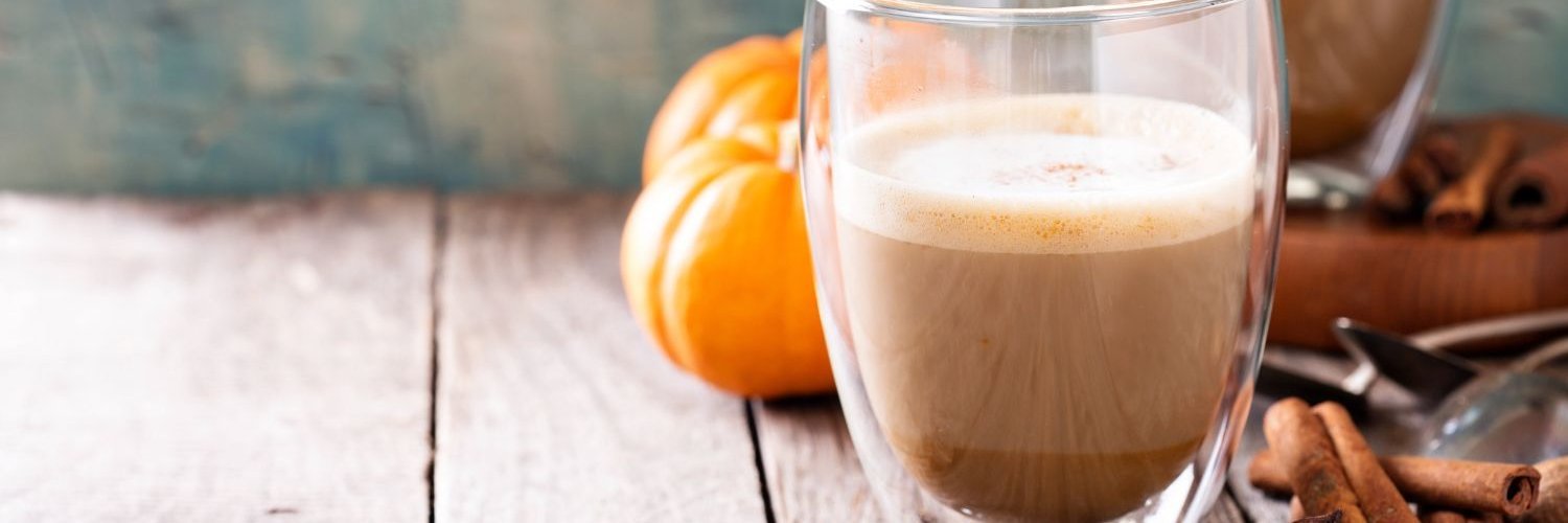 Ein Glas Pumpkin Spice Latte mit etwas Milchschaum steht vor kleinen Kürbissen auf einem Holztisch. Neben dem Glas liegen Zimtstangen und Sternanis.