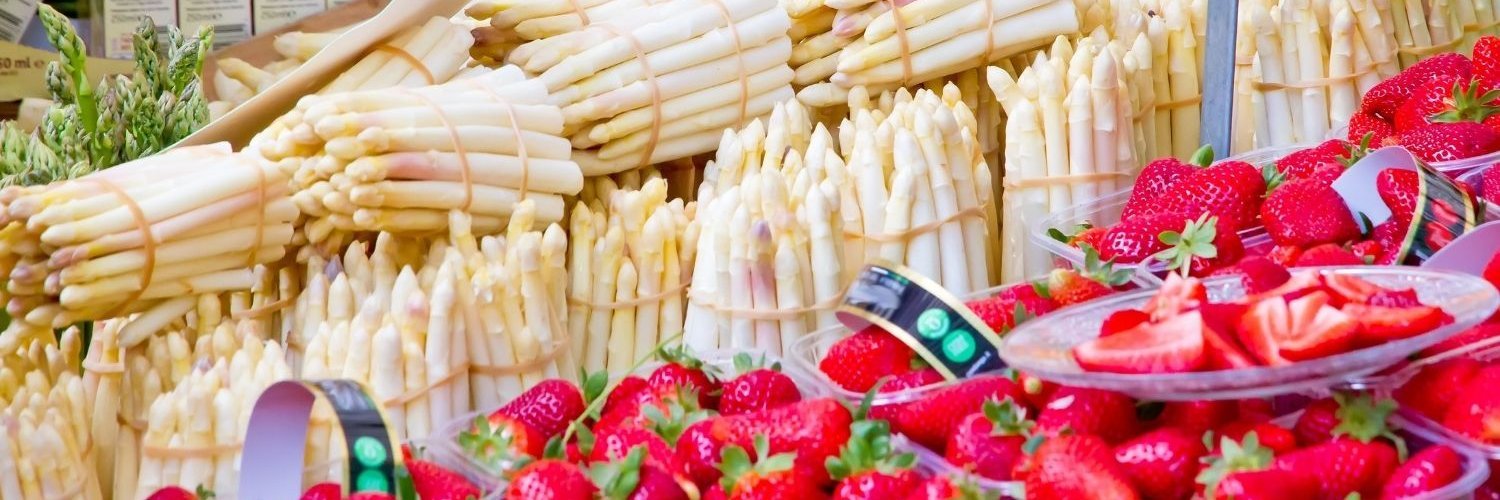 An einem Marktstand werden viele Bündel mit weißem Spargel und Körbchen mit Erdbeeren angeboten.
