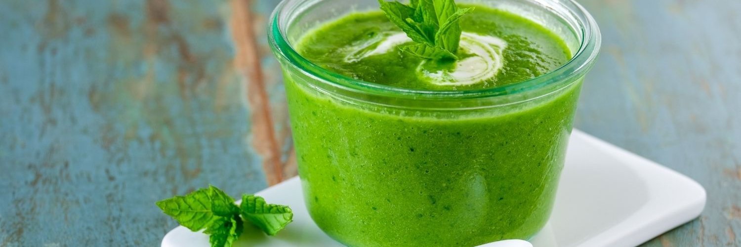 Auf einem weißen Brett steht eine Glasschüssel mit grüner Erbsensuppe, garniert mit Minzblättern. Daneben liegt ein Löffel.