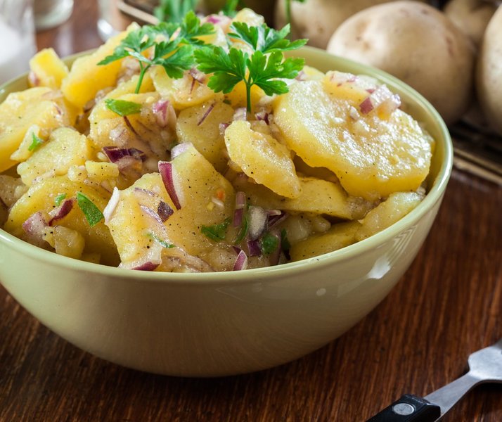 Eine Schale mit Kartoffelsalat mit roten Zwiebeln und Petersiliengarnitur steht auf dem Tisch.
