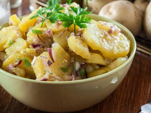 Eine Schale mit Kartoffelsalat mit roten Zwiebeln und Petersiliengarnitur steht auf dem Tisch.