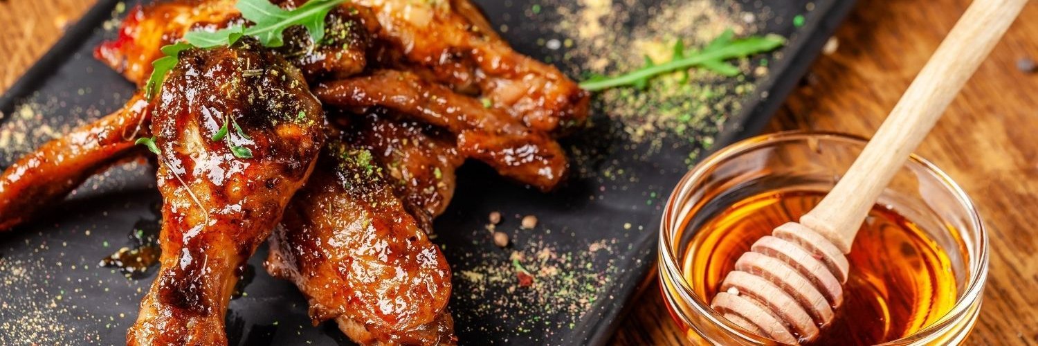 Auf einer Schieferplatte liegen glasierte Hähnchenflügel, daneben steht eine Glasschüssel mit Honig und einem Honiglöffel darin.