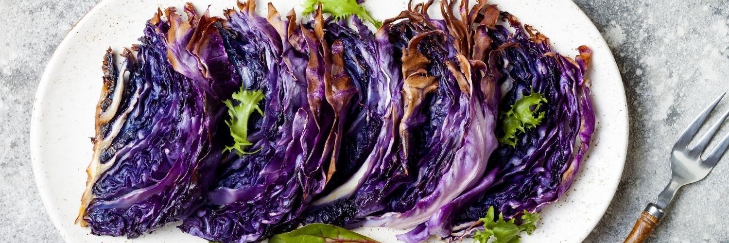 Auf einer ovalen weißen Platte liegen gegarte halbierte Rotkohlscheiben garniert mit Salatblättchen, daneben eine Gabel mit Holzgriff.
