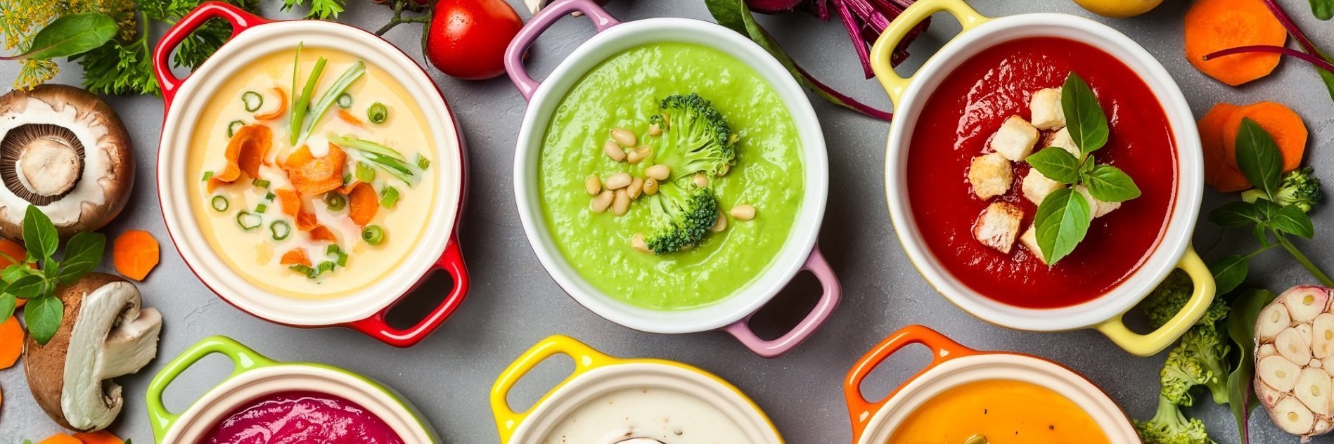 Ein Tisch mit verschiedenen Suppen mit Brokkoli, Rote Bete, Möhre, Champignon, drumherum pflanzliche Zutaten, wie Kartoffeln, Tomate, Lauchzwiebeln, Petersilie und mehr ist abgebildet.