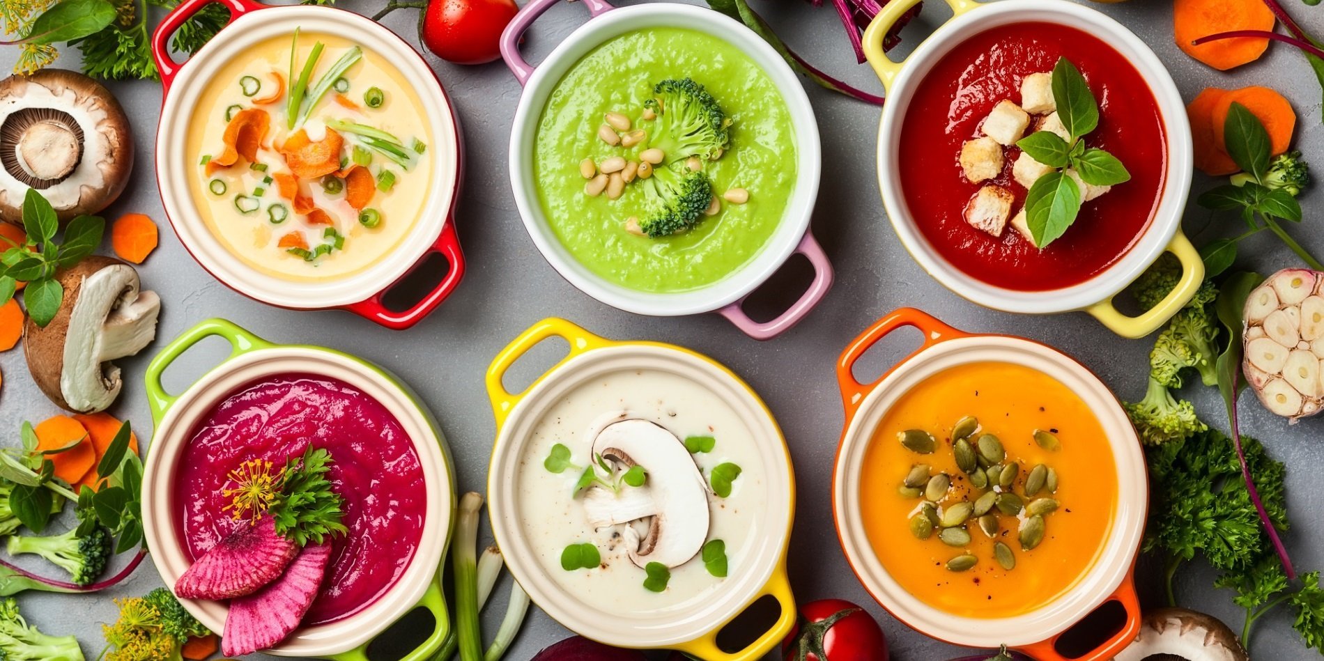 Ein Tisch mit verschiedenen Suppen mit Brokkoli, Rote Bete, Möhre, Champignon, drumherum pflanzliche Zutaten, wie Kartoffeln, Tomate, Lauchzwiebeln, Petersilie und mehr ist abgebildet.. Artikel "Vegane Ernährung" öffnen.