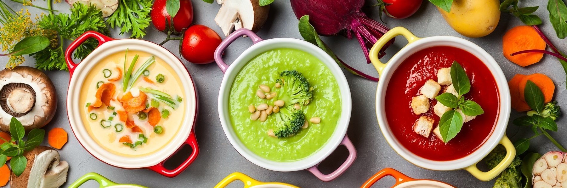 Ein Tisch mit verschiedenen Suppen mit Brokkoli, Rote Bete, Möhre, Champignon, drumherum pflanzliche Zutaten, wie Kartoffeln, Tomate, Lauchzwiebeln, Petersilie und mehr ist abgebildet.