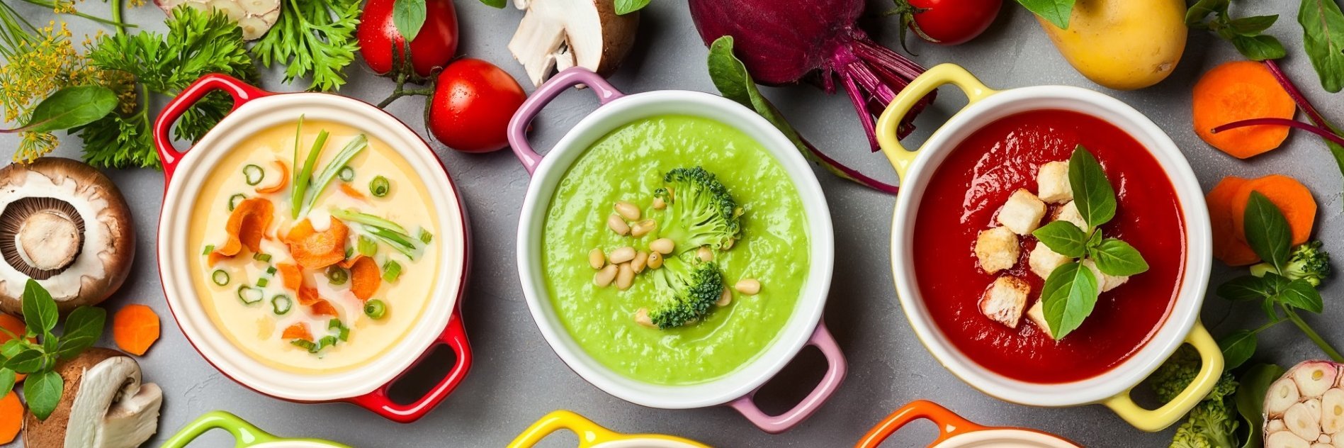Ein Tisch mit verschiedenen Suppen mit Brokkoli, Rote Bete, Möhre, Champignon, drumherum pflanzliche Zutaten, wie Kartoffeln, Tomate, Lauchzwiebeln, Petersilie und mehr ist abgebildet.