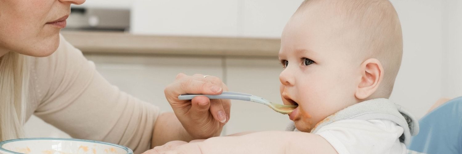 Eine Frau füttert ein Baby im Hochstuhl mit Brei und einem Löffel.