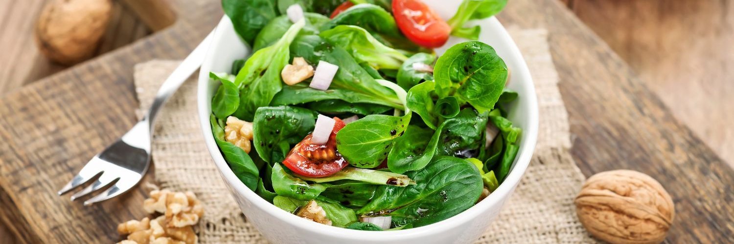 Auf einem Holzbrett steht eine weiße Schüssel mit Feldsalat, Walnüssen, Käsewürfeln und Tomate. Daneben liegen halbierte Walnüsse und eine Gabel.