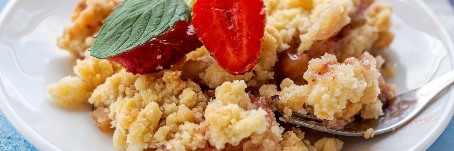 Auf einem hellblauen Untergrund steht ein weißer Teller mit Rhabarber-Crumble, dekoriert mit halbierten Erdbeeren und Melisse. 