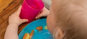 Kleinkind trinkt aus pinkem Becher, während sich in der Auffangschale seines blauen Gummilätzchens Karotten und Kartoffeln sammeln.