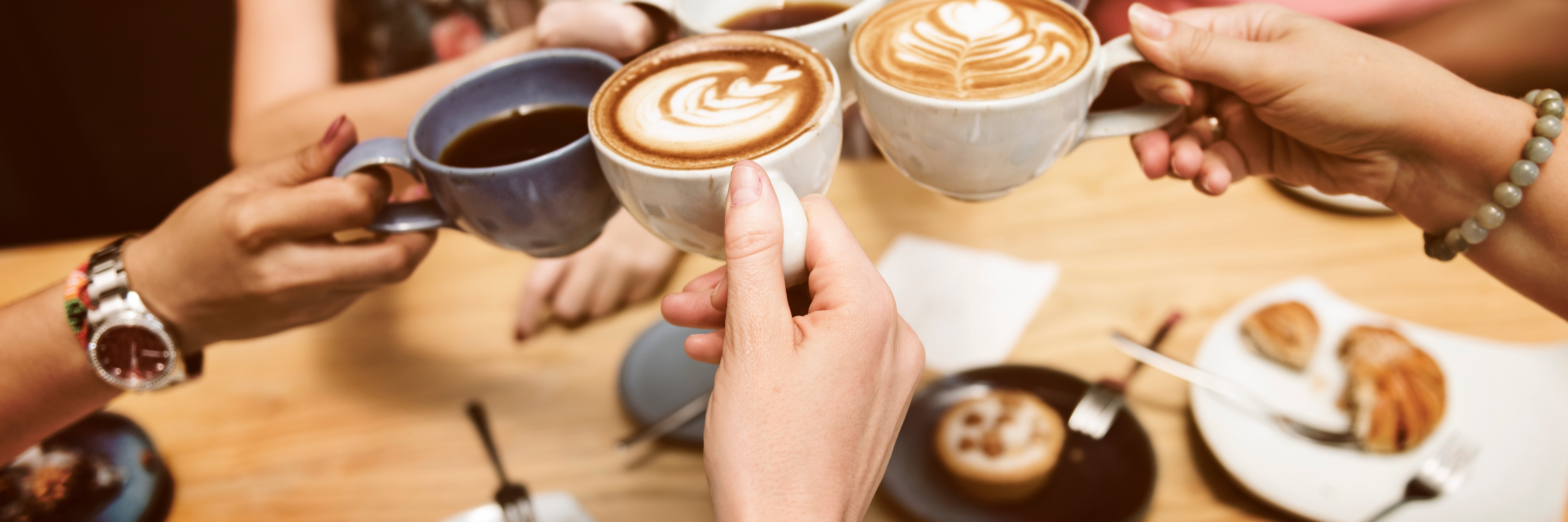 Fünf Frauen unterschiedlicher Hautfarbe stoßen mit ihren Tassen miteinander an. In den Tassen sind verschiedene Kaffeevarianten.