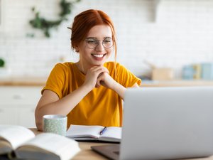 Junge Frau mit Tasse und Buch vor sich schaut auf Laptop.