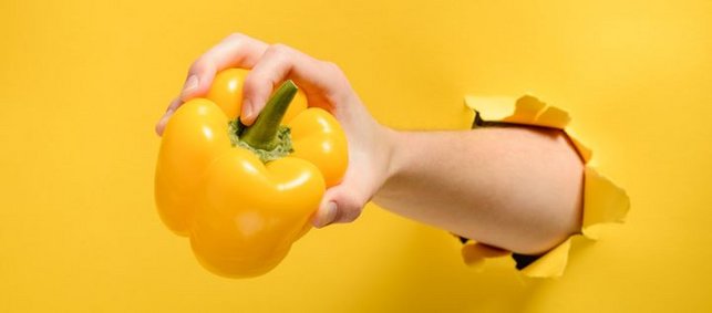 gelbe Paprika in einer Hand, die mit Arm durch eine gelbe Wand greift
