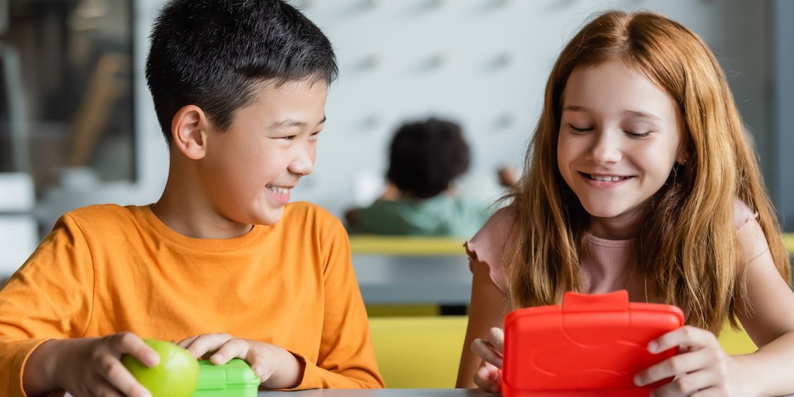 Zwei offensichtlich gut gelaunte Schulkinder öffnen ihre Brotdosen.