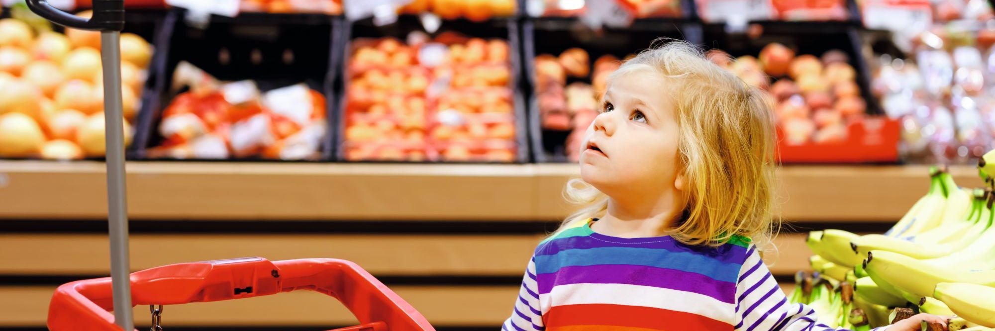 Ein Kitakind steht neben einem Einkaufswagen in der Obst- und Gemüseabteilung eines Supermarktes.
