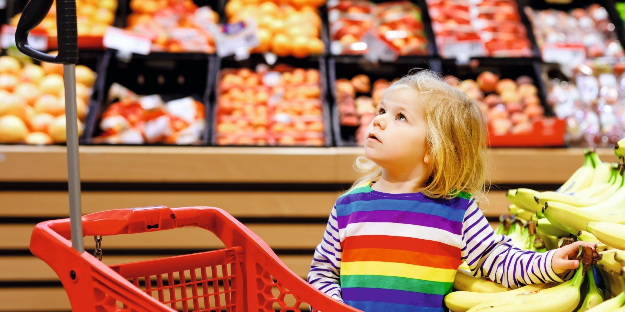 Ein Kitakind steht neben einem Einkaufswagen in der Obst- und Gemüseabteilung eines Supermarktes. . Artikel "Einkaufen mit KiTa-Kindern" öffnen.