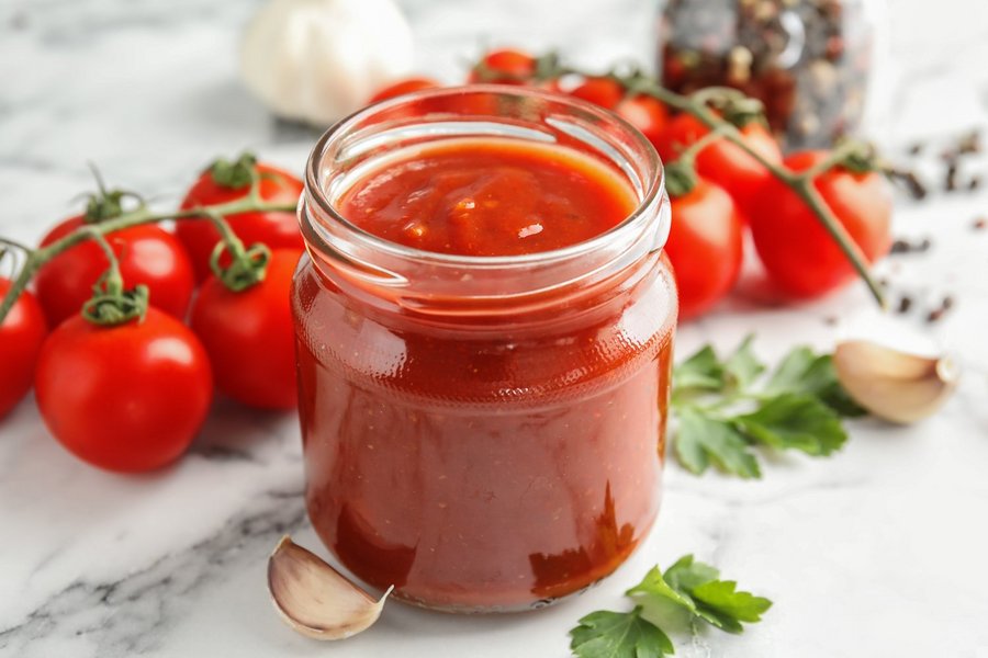Im Vordergrund ein offenes Glas mit Tomatensoße. Dahinter frische Tomaten und Gewürze.