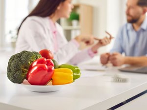 Eine Ernährungsberaterin berät einen jungen Mann. Im Vordergrund ist eine Teller gefüllt mit frischem Gemüse.